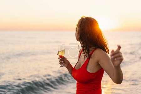 A cheerful woman in a vibrant red swimsuit stands by the seaside, holding a glass of white wine. The setting sun casts a warm golden hue over the scene, highlighting her joyous expression.