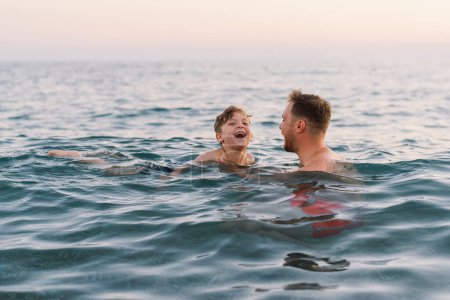 Un père et son fils partagent un moment agréable tout en nageant dans les eaux calmes de l'océan alors que la lumière du soir s'estompe doucement en arrière-plan. Leurs sourires traduisent un sentiment de plaisir. Bonne fête des Pères