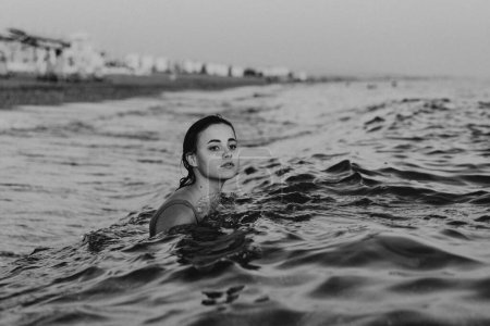 Téléchargez les photos : Une jeune femme souriante nage dans les eaux calmes de l'océan en début de soirée. Sa tête et ses épaules sont visibles au-dessus de la ligne de flottaison - en image libre de droit