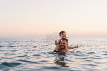 Un père et son fils partagent un moment agréable tout en nageant dans les eaux calmes de l'océan alors que la lumière du soir s'estompe doucement en arrière-plan. Leurs sourires traduisent un sentiment de plaisir. Bonne fête des Pères
