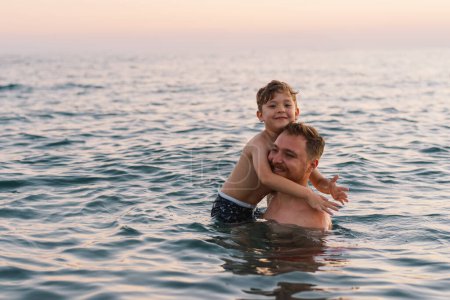Ein Vater und sein Sohn verbringen einen angenehmen Moment beim Schwimmen im ruhigen Ozeanwasser, während das Abendlicht im Hintergrund sanft verblasst. Ihr Lächeln vermittelt ein Gefühl der Freude. Glücklicher Vatertag