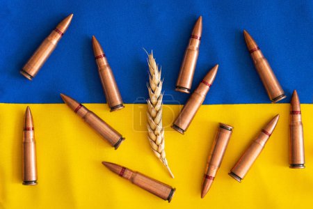 Wheat grains with yellow and blue Ukrainian flag on wooden background with several metal bullet shells. The concept of the photo suggests that grain can be used as a weapon. Hunger as a weapon