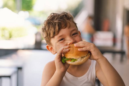 Niño comiendo sándwich y papas fritas en la mesa. Aparece enfocado en su comida, con un sándwich en una mano y papas fritas en la otra. Un chico come comida rápida al aire libre..