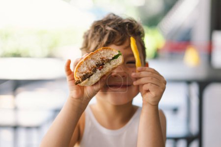 Kleiner Junge isst Sandwich und Pommes am Tisch. Er wirkt konzentriert auf seine Mahlzeit, mit einem Sandwich in der einen und Pommes in der anderen Hand. Ein Junge isst Fast Food im Freien.