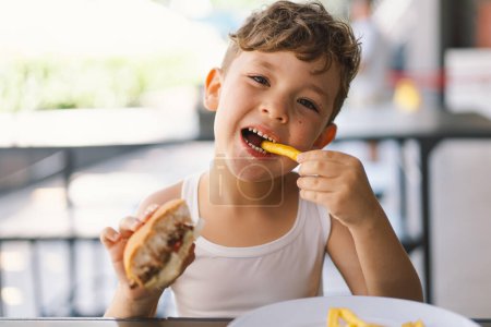 Photo for Little Boy Eating Sandwich and French Fries at Table. He appears focused on his meal, with a sandwich in one hand and a French Fries in the other hand. A boy eats fast food outdoors. - Royalty Free Image