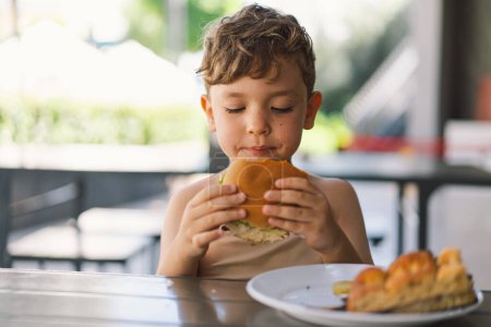 Kleiner Junge isst Sandwich und Pommes am Tisch. Er wirkt konzentriert auf seine Mahlzeit, mit einem Sandwich in der einen und Pommes in der anderen Hand. Ein Junge isst Fast Food im Freien.