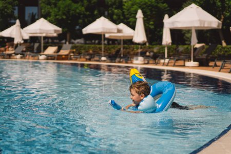 Ein Junge mit freudigem Gesichtsausdruck schwebt an einem sonnigen Tag in einem Schwimmbad und benutzt einen Schwimmkreis, der wie ein blauer Vogel aussieht..