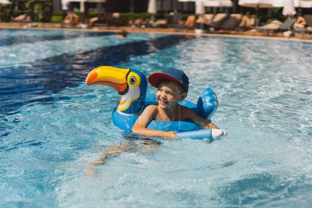 A boy with a joyful expression is floating in a swimming pool on a sunny day, using an swimming circle designed to look like a blue bird.