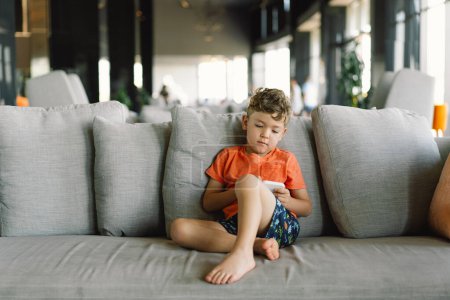 Ein Junge mit lockigem Haar liegt gemütlich auf einem grauen Sofa mit Kissen und ist am Telefon tief in das Spiel vertieft. gemütlicher Tag im Haus.