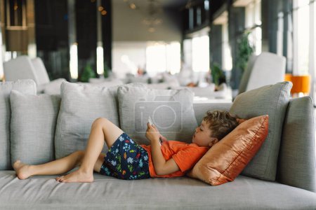 Téléchargez les photos : Un garçon aux cheveux bouclés s'appuie confortablement sur un canapé gris avec des oreillers, profondément immergé dans le jeu au téléphone. journée tranquillement à l'intérieur. - en image libre de droit