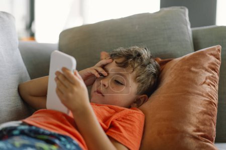 Un garçon aux cheveux bouclés s'appuie confortablement sur un canapé gris avec des oreillers, profondément immergé dans le jeu au téléphone. journée tranquillement à l'intérieur.