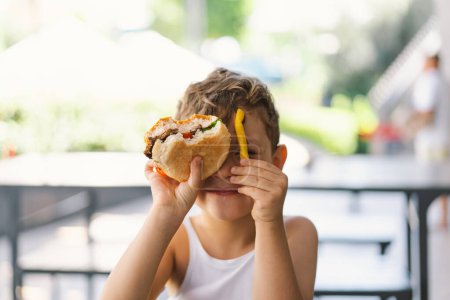 Photo for Little Boy Eating Sandwich and French Fries at Table. He appears focused on his meal, with a sandwich in one hand and a French Fries in the other hand. A boy eats fast food outdoors. - Royalty Free Image