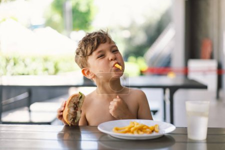 Niño comiendo sándwich y papas fritas en la mesa. Aparece enfocado en su comida, con un sándwich en una mano y papas fritas en la otra. Un chico come comida rápida al aire libre..