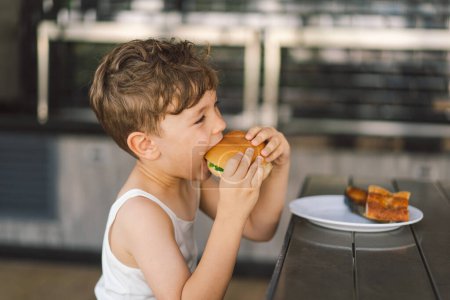 Little Boy Eating Sandwich and French Fries at Table. He appears focused on his meal, with a sandwich in one hand and a French Fries in the other hand. A boy eats fast food outdoors.