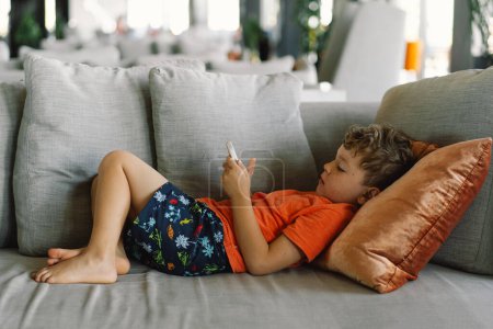 Ein Junge mit lockigem Haar liegt gemütlich auf einem grauen Sofa mit Kissen und ist am Telefon tief in das Spiel vertieft. gemütlicher Tag im Haus.