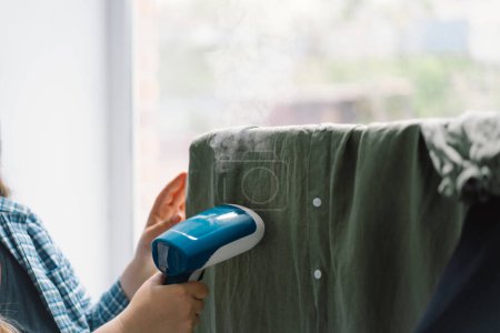 Femme utilisant un vapeur portable bleu sur un vêtement vert à la maison pendant la journée. Lumière naturelle du jour qui éclaire la texture des tissus et la vapeur en action.