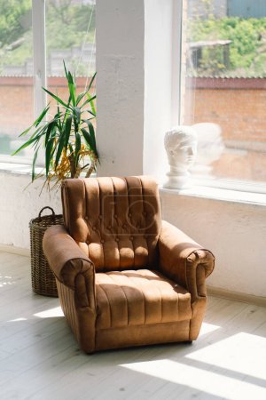 Un sillón de cuero vintage toma el sol cerca de una gran ventana con una vista despejada en el exterior, acompañado de una planta en maceta verde y una canasta de mimbre en una habitación serena y luminosa..