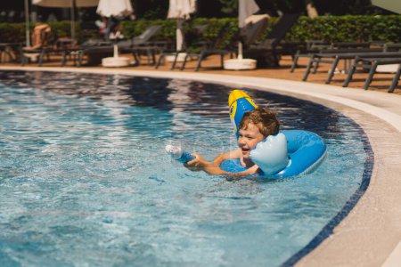 Un garçon avec une expression joyeuse flotte dans une piscine par une journée ensoleillée, en utilisant un cercle de natation conçu pour ressembler à un oiseau bleu.
