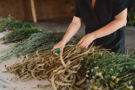 A woman stands at a wooden table in a rustic, diligently working on assembling a natural wreath from an assortment of freshly cut branches and foliage that are scattered across the work surface.