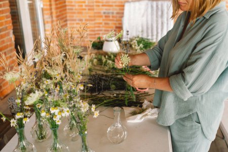 A woman stands at a wooden table, artfully arranging assorted green plants and branches in a spacious workshop. She appears concentrated on selecting the perfect placement for each plant