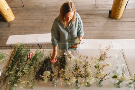 A woman stands at a wooden table, artfully arranging assorted green plants and branches in a spacious workshop. She appears concentrated on selecting the perfect placement for each plant