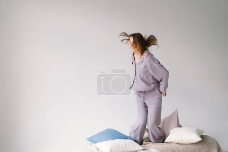 A cheerful woman tosses a pillow with a joyous expression. She stands barefoot on a beige blanket with a stack of pillows and soft textiles around her, suggesting a comfortable, lighthearted setting
