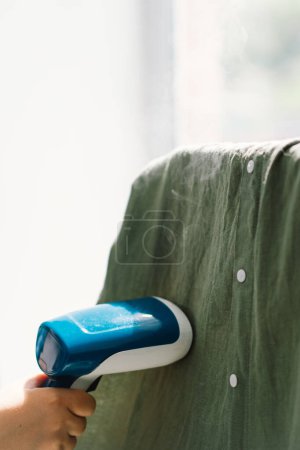 Femme utilisant un vapeur portable bleu sur un vêtement vert à la maison pendant la journée. Lumière naturelle du jour qui éclaire la texture des tissus et la vapeur en action.