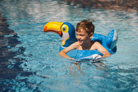 A boy with a joyful expression is floating in a swimming pool on a sunny day, using an swimming circle designed to look like a blue bird.