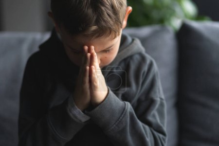 Un jeune garçon est assis sur un canapé doux, les mains serrées devant lui comme s'il priait ou méditait. Religion et concept de foi.