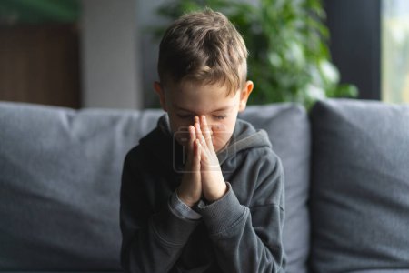 A young boy is seated on a soft sofa, his hands pressed together in front of him as if in is praying or meditation. Religion and faith concept.