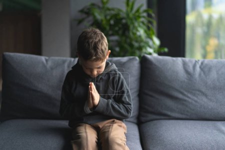 A young boy is seated on a soft sofa, his hands pressed together in front of him as if in is praying or meditation. Religion and faith concept.