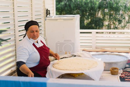 Eine Frau bereitet ein großes, dünnes Stück Fladenbrot zu. Sie arbeitet an einem Marktstand im Freien, umgeben von Kochutensilien und Zutaten, die einen Einblick in lokale kulinarische Praktiken geben..
