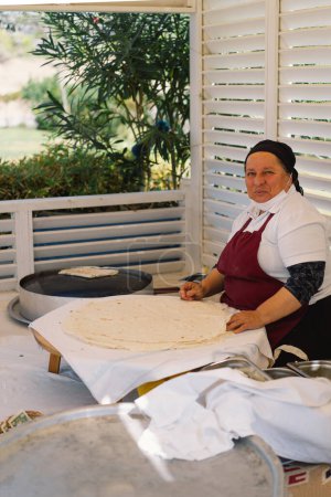 Eine Frau bereitet ein großes, dünnes Stück Fladenbrot zu. Sie arbeitet an einem Marktstand im Freien, umgeben von Kochutensilien und Zutaten, die einen Einblick in lokale kulinarische Praktiken geben..