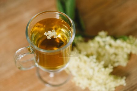 Una taza de vidrio transparente llena de té de flor de saúco se coloca en una mesa de madera rústica. Flores de saúco frescas y una tabla de cortar de madera están cerca, creando un ambiente acogedor y natural.