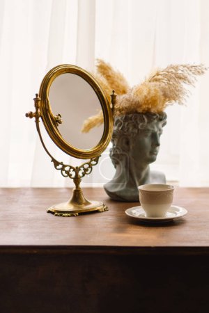 Gold-framed mirror stands on a wooden table, beside a classical bust vase adorned with pampas grass. A simple white cup of coffee completes the calming and sophisticated setting.