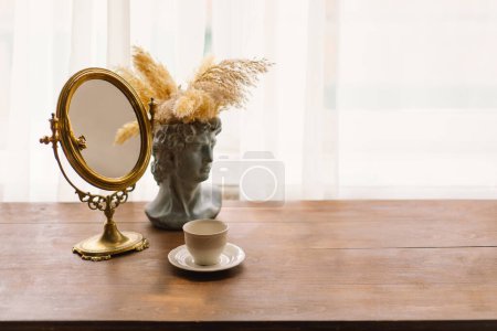 Auf einem Holztisch steht ein goldgerahmter Spiegel neben einer klassischen Büstenvase, die mit Pampasgras geschmückt ist. Eine einfache weiße Tasse Kaffee rundet das beruhigende und raffinierte Ambiente ab.