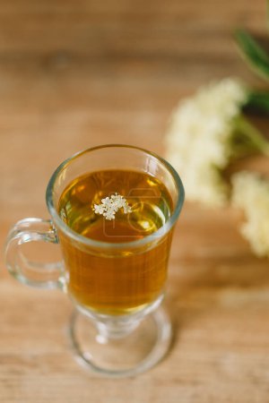 A clear glass mug filled with elderflower tea is placed on a rustic wooden table. Fresh elderflowers and a wooden cutting board are nearby, creating a cozy and natural atmosphere.
