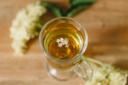 Une tasse en verre transparent remplie de thé de sureau est placée sur une table en bois rustique. Des fleurs de sureau fraîches et une planche à découper en bois sont à proximité, créant une atmosphère chaleureuse et naturelle.