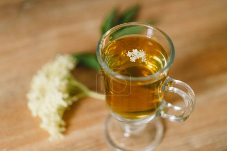A clear glass mug filled with elderflower tea is placed on a rustic wooden table. Fresh elderflowers and a wooden cutting board are nearby, creating a cozy and natural atmosphere.
