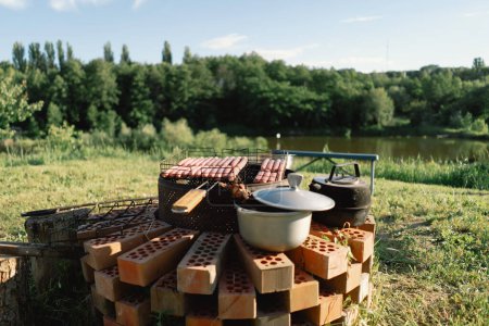 Barbecue set up on a sunny day near a river. The grill is filled with meat, and a kettle sits on a brick base beside it. Smoke rises from the grill, filling the air with the aroma of grilling food.