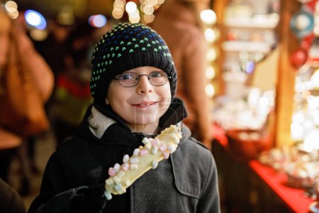 Photo for Little kid boy, cute child eating bananas covered with chocolate, marshmellows and colorful sprinkles near sweet stand with gingerbread and nuts. Happy boy on Christmas market in Germany - Royalty Free Image
