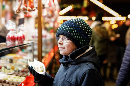 Photo for Little kid boy, cute child eating bananas covered with chocolate, marshmellows and colorful sprinkles near sweet stand with gingerbread and nuts. Happy boy on Christmas market in Germany - Royalty Free Image