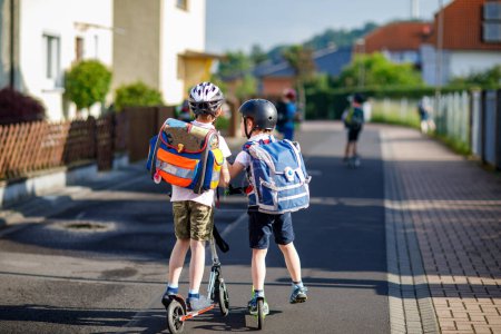 Zwei Schulkinder mit Schutzhelm fahren an einem sonnigen Tag mit dem Roller in der Stadt mit dem Rucksack. Glückliche Kinder in bunten Kleidern radeln auf dem Schulweg. Sicherer Schulweg für Kinder im Freien.