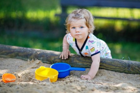 Téléchargez les photos : Petite fille mignonne jouant dans le sable sur une aire de jeux extérieure. Beau bébé en vêtements d'été s'amusant par une chaude journée d'été ensoleillée. Activité de plein air pour les tout-petits - en image libre de droit