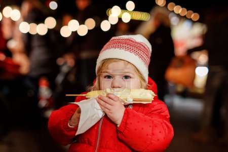 Photo for Little baby girl, cute child eating bananas covered with chocolate, marshmellows and colorful sprinkles near sweet stand with gingerbread and nuts. Happy toddler on Christmas market in Germany - Royalty Free Image