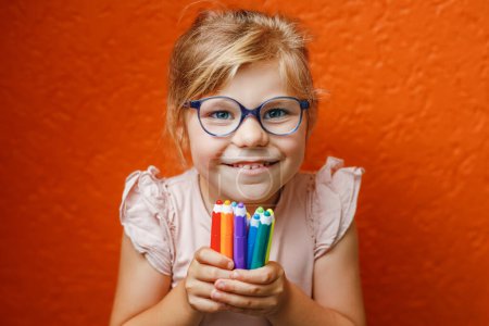Téléchargez les photos : Happy cute little preschooler girl with glasses holding colorful pencils and making gesture while looking at camera. Playful child with pencils. Imagination and creativity at school concept - en image libre de droit