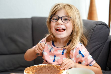 Foto de Adorable little girl having breakfast, eating pancakes with chocolate cream. Preschool child smiling. Sweet food for children - Imagen libre de derechos