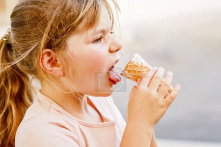 Foto de Pequeña niña preescolar adorable comiendo helado en cono de gofre en el día soleado de verano. Feliz niño pequeño come postre helado. Comida dulce en días calurosos de verano. - Imagen libre de derechos