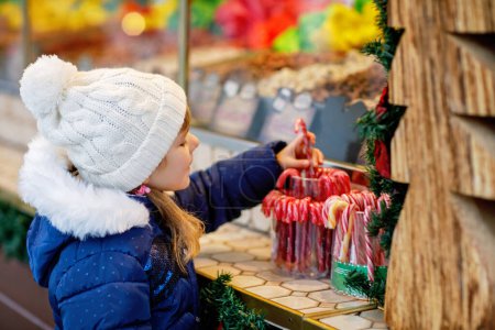 Foto de Pequeña niña linda preescolar comprar bastón de caramelo de un puesto de dulces en el mercado de Navidad. Niño feliz en el mercado familiar tradicional en Alemania. Preescolar en ropa de invierno colorida. - Imagen libre de derechos