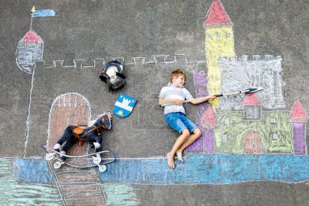 Photo for Little active kid boy drawing knight castle and fortress with colorful chalks on asphalt. Happy child in helmet and with spear and rocking horse toy having fun with playing knight game and painting. - Royalty Free Image